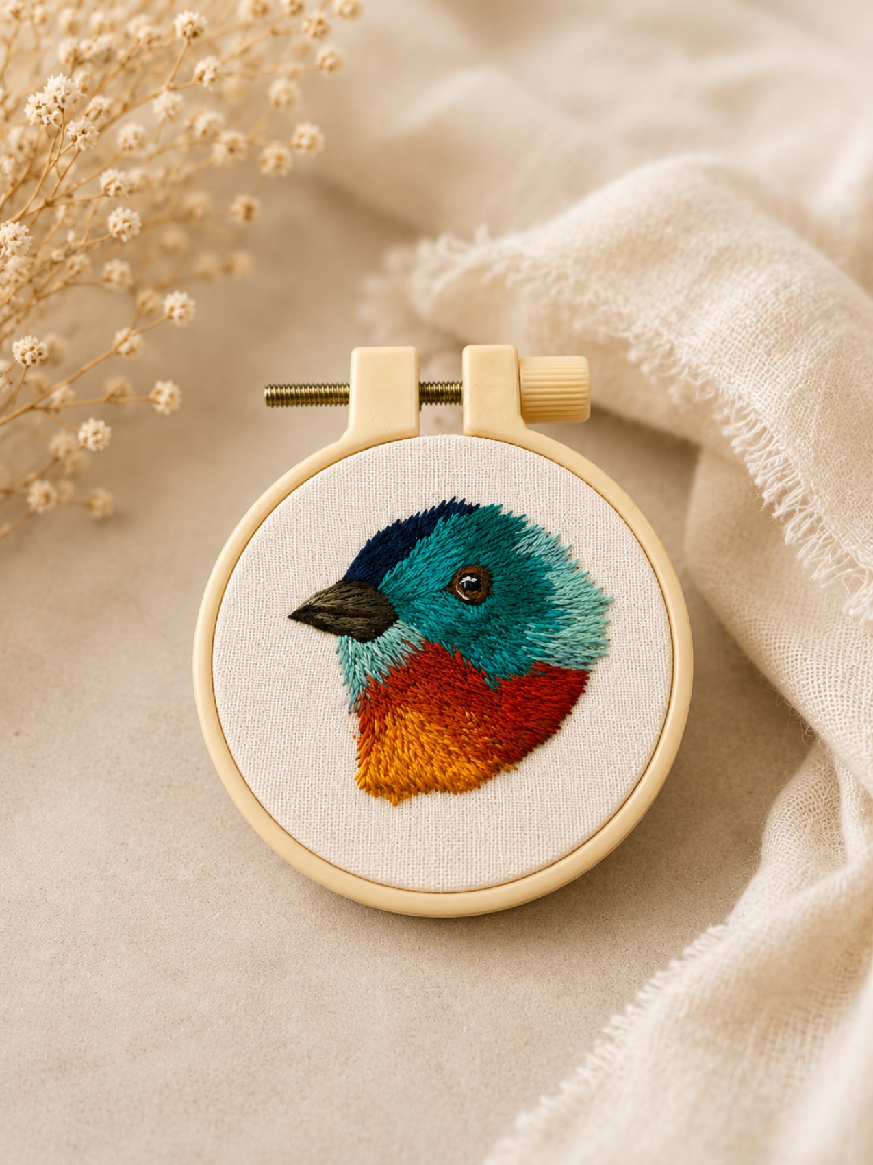 A painted bunting, hand-embroidered with a teal-blue head, red throat and orange belly on linen.