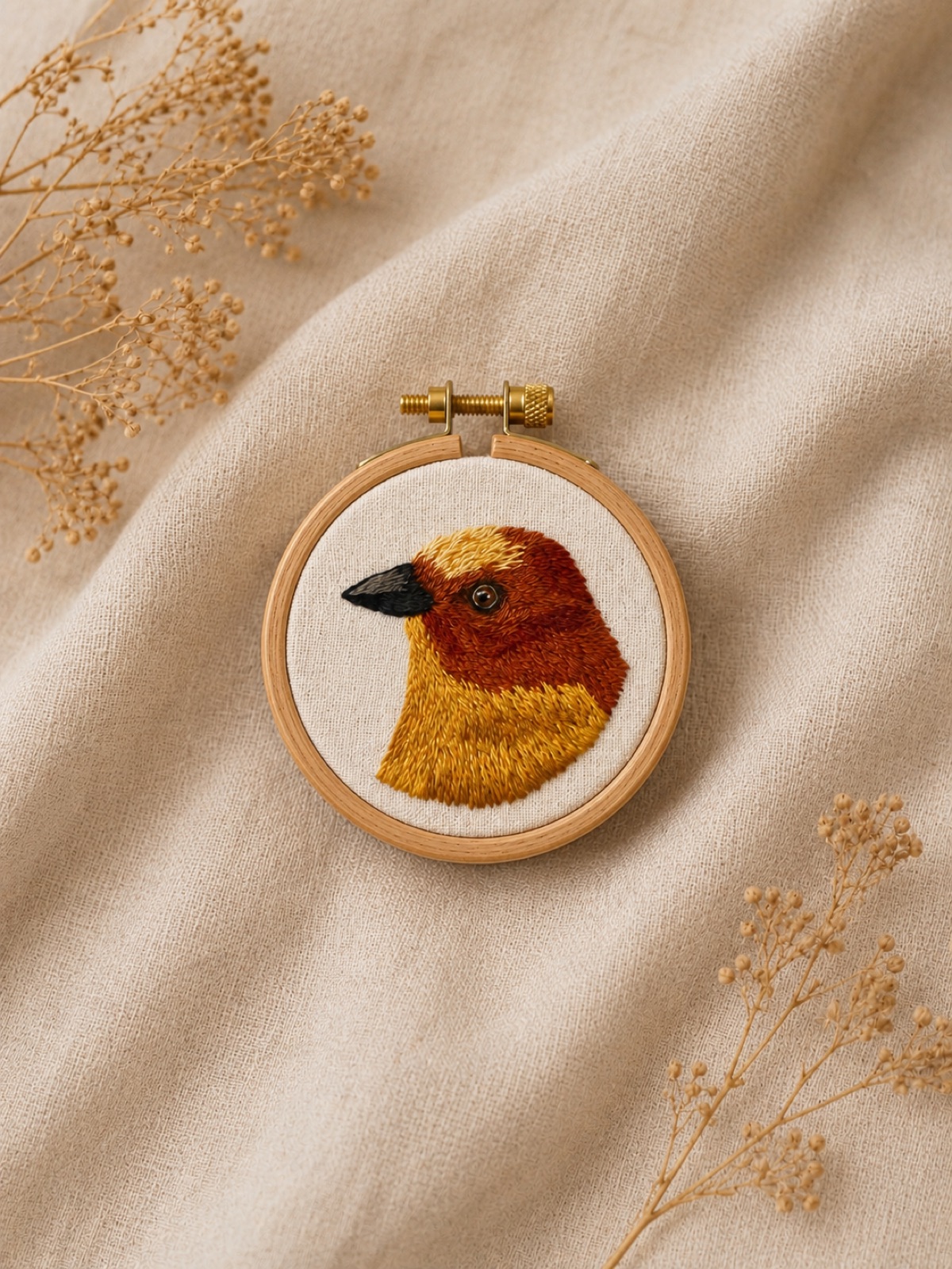 A weaver bird in profile, hand-embroidered in warm amber and chestnut thread with a black beak, on raw cotton.